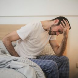 A man holding his forehead while sitting on the edge of his bed.